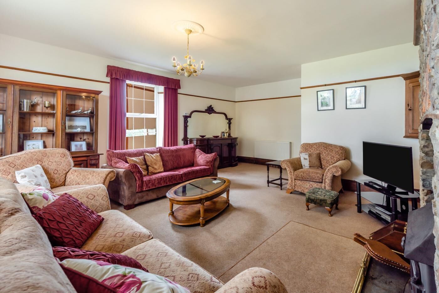 A spacious living room with a large sofa, a smaller sofa, a coffee table, a television, and a fireplace. There is a wooden cabinet with glass doors and a chandelier.