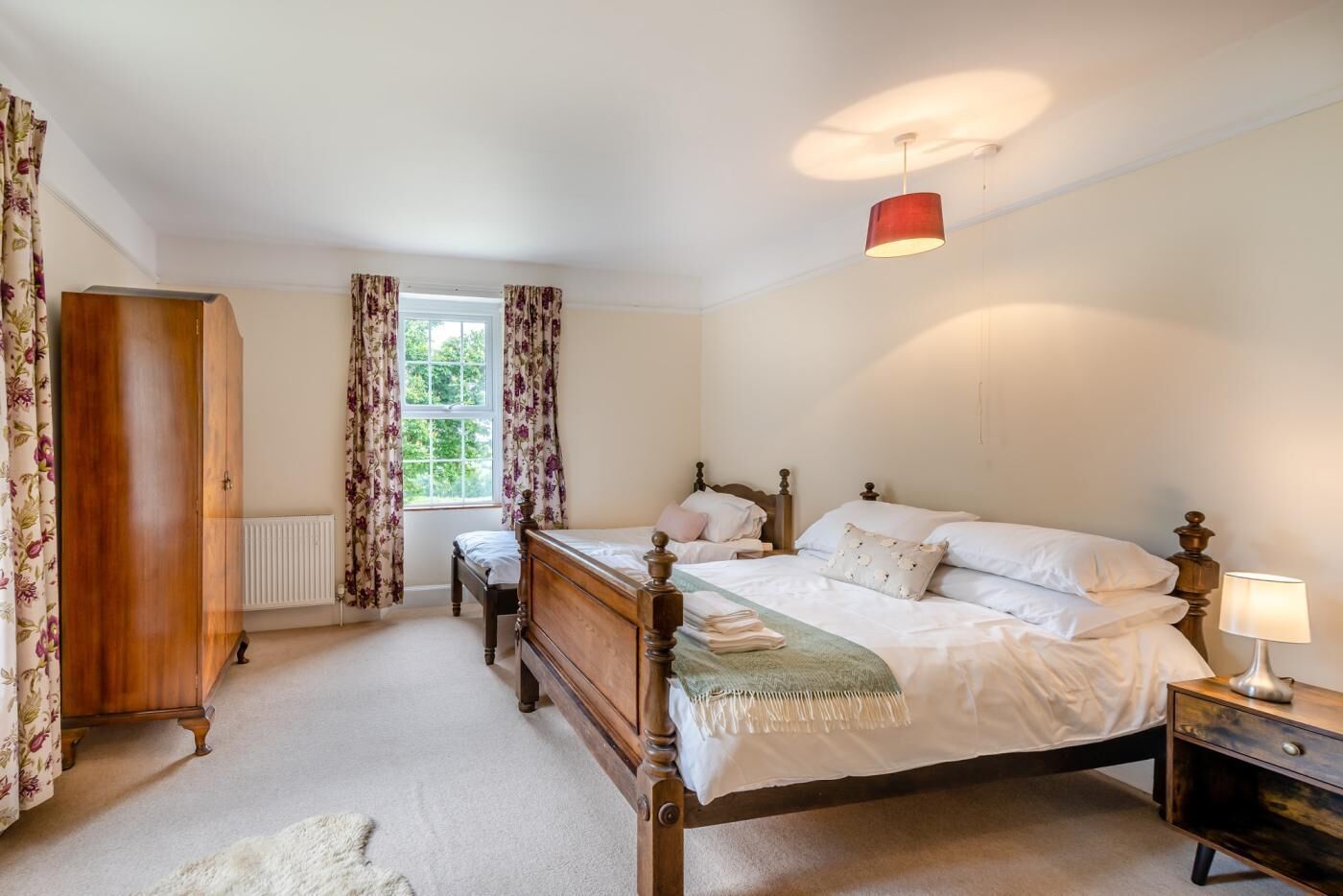 Une chambre à coucher avec un lit en bois, une table de chevet, une lampe rouge et une fenêtre ornée de rideaux à motifs floraux.