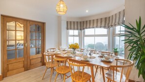 A dining area with a wooden table set for six, surrounded by wooden chairs. There is a large window with a view of the outdoors, a hanging light fixture, and a door with glass panels. - 5 Bed in Mynytho (oc-c28916) (Mynytho)