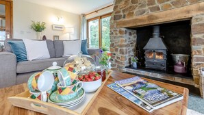 Ein gemütliches Wohnzimmer mit Kamin aus Stein, einer Couch, einem Couchtisch mit Geschirr und Zeitschriften sowie einem Fenster mit Blick ins Grüne.