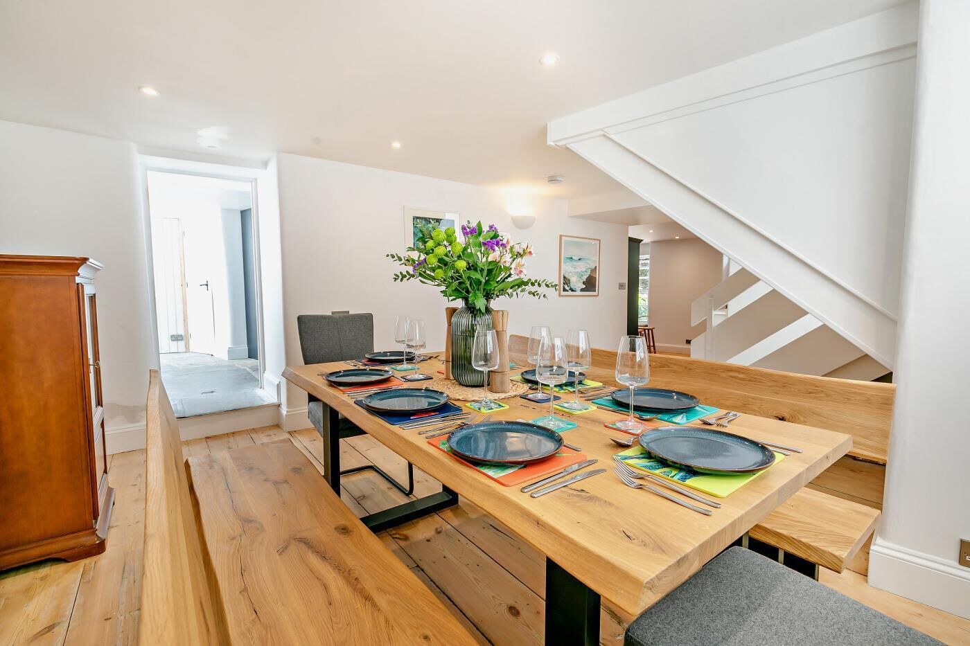 Un coin repas avec une table en bois dressée pour six personnes, un vase rempli de fleurs et un escalier en arrière-plan.
