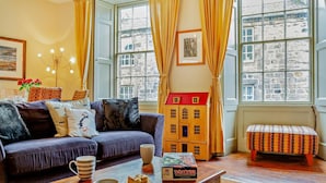A cozy living room with a sofa, a wooden playhouse, a coffee table with mugs and a board game, and large windows with curtains. - 2 Bed in Alnwick (oc-f28915) (Alnwick)