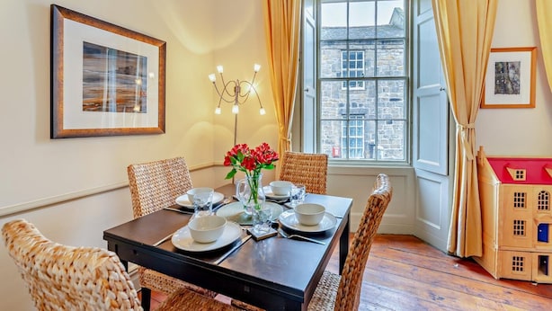 A dining area with a table set for four, a vase with red flowers, and a view of a stone building through a window with curtains. - 2 Bed in Alnwick (oc-f28915) (Alnwick)
