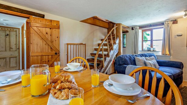 A dining area with a wooden table set for breakfast, including croissants and orange juice, and a staircase leading to an upper level. - 3 Bed in Mortehoe (oc-s29000) (Woolacombe)