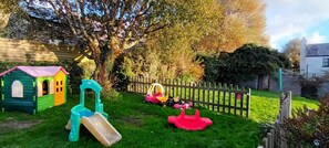 Ein eingezäunter Garten mit einem grünen Spielhäuschen, einer Rutsche und einer Schaukel.
