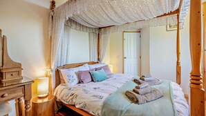 A four-poster canopy bed with a floral bedspread, a bedside table with a lamp, and a wooden nightstand. - 3 Bed in Mortehoe (oc-s29000) (Woolacombe)