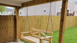 Una silla mecedora de madera colgada por cadenas, un suelo de hormigón y una valla de madera.