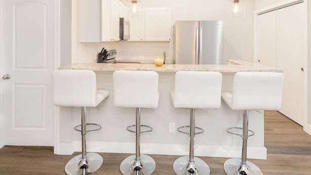 Una cocina moderna con una isla central, cuatro taburetes blancos tipo bar, electrodomésticos de acero inoxidable e iluminación colgante.