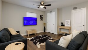 Una sala moderna con un televisor de pantalla plana, dos sofás negros, una mesa de centro de madera, un tapete de piel de vaca y un ventilador de techo.