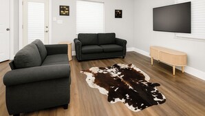 Una sala de estar con dos sofás, piso de madera, alfombra de cuero de vaca, televisor y un banco.