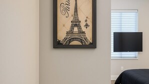 Un dormitorio con un cuadro enmarcado de la Torre Eiffel, un televisor de pantalla plana montado en la pared y una cama con cubrecolor negro.
