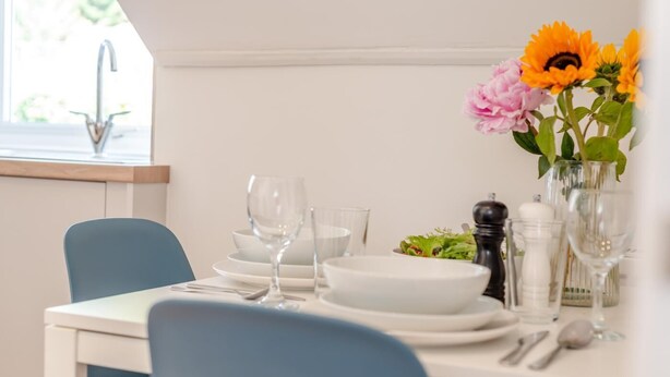 A dining table set with plates, glasses, and cutlery, a vase with flowers, and a window with a view. - 1 Bed in Looe (oc-s29019) (Looe)