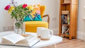 A room with a yellow sofa, a bookshelf, a white table with a book and a mug, and a vase with flowers. - 1 Bed in Looe (oc-s29019) (Looe)