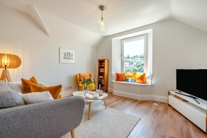 A modern living room with a sofa, a coffee table, a bookshelf, a window seat with cushions, and a television.