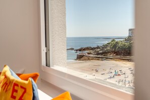 A window view of a beach with a rocky shoreline, a pier, and a building in the background.