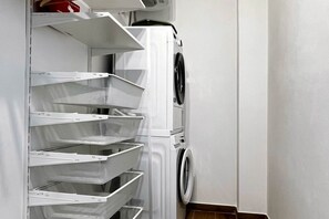 A white washing machine with a front-loading door, adjacent to a wall-mounted drying rack with multiple mesh shelves. - Alma Suites - Terrace Penthouse (Firenze)