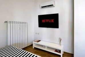 Ein modernes Zimmer mit einem weißen Heizkörper, einer weißen Bank mit Schachbrett und einem schwarzen, an der Wand befestigten Fernseher, der 'NETFLIX' anzeigt.