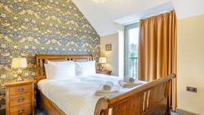 Ein Schlafzimmer mit einem hölzernen Bett, zwei Nachttischen mit Lampen, blumigem Wandpapier und Blick durch ein Fenster mit Vorhängen.
