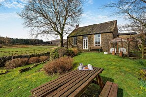 A stone house with a large window, a wooden bench, and a picnic table with food. - 2 Bed in Grisedale (oc-c29159) (Grisdale)