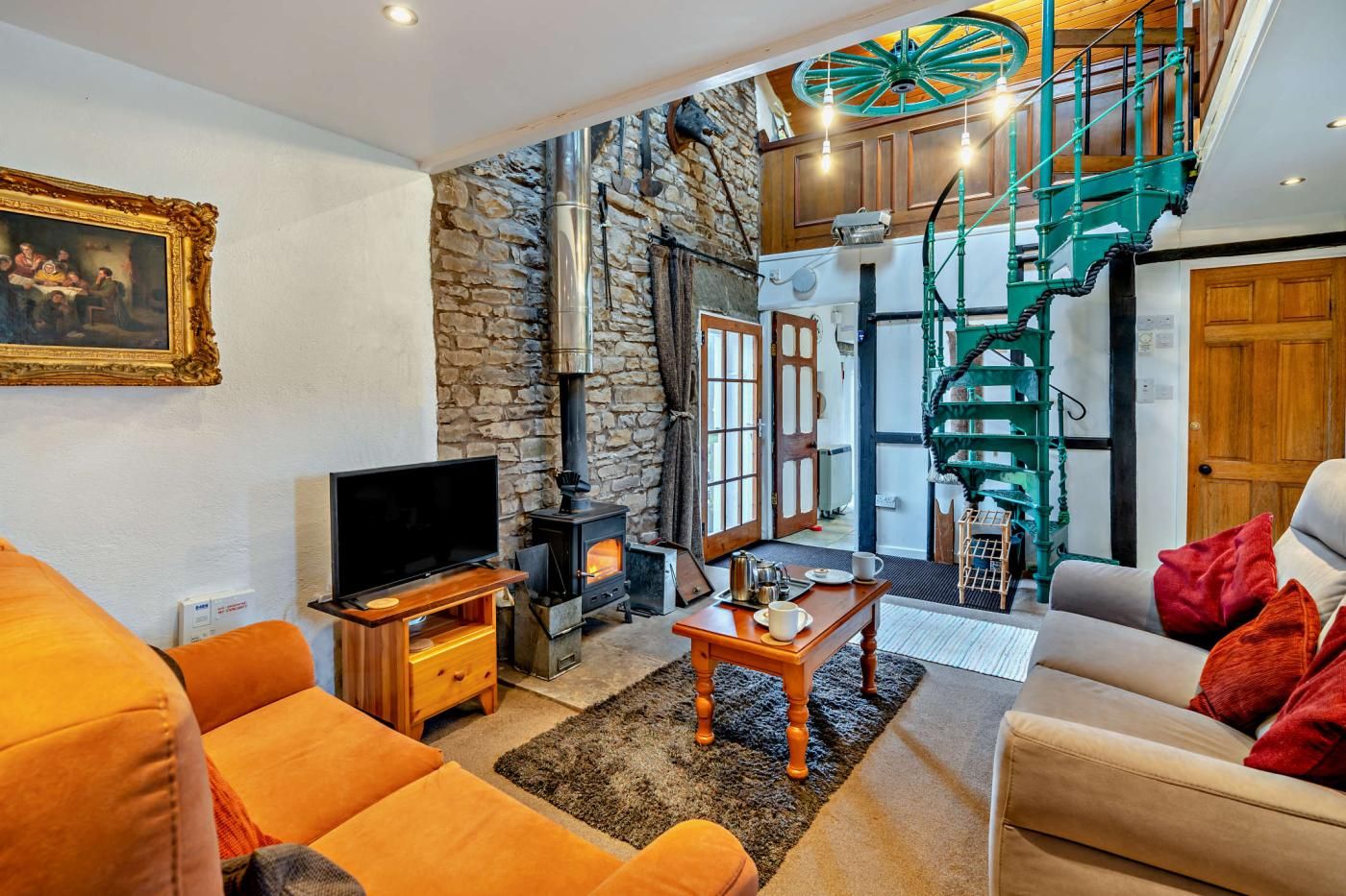 A cozy living room with a stone wall, a fireplace, a television, and a staircase with a green railing.
