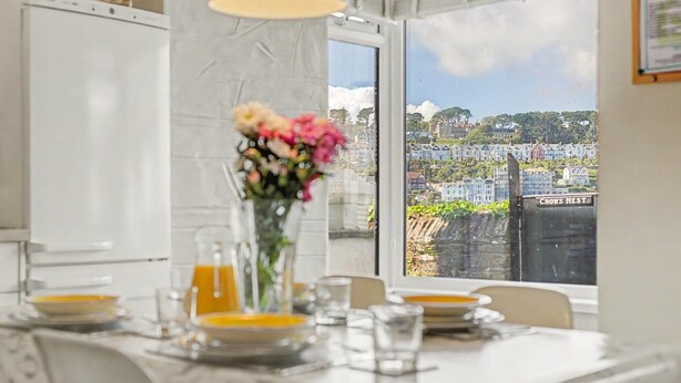 A dining table with a vase of flowers, a white refrigerator, and a view of a residential area through a window. - 2 Bed in Polruan (oc-denzil) (Fowey)