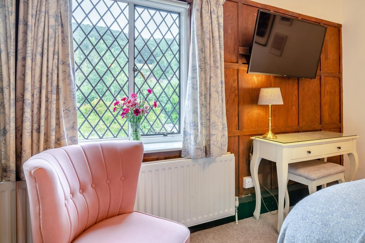 Ein Zimmer mit einem rosa Stuhl, einem Holztisch mit Lampe, einem an der Wand montierten Fernseher und einem Fenster mit Vorhängen und einem Blumenstrauß.