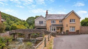 A two-story house with a stone bridge over a stream, surrounded by greenery. - 1 Bed in Allerford (oc-m29128) (Allerford)