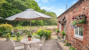 A patio with a round table and chairs, a large umbrella, and a brick building with hanging flower baskets. - 1 Bed in Allerford (oc-m29128) (Allerford)