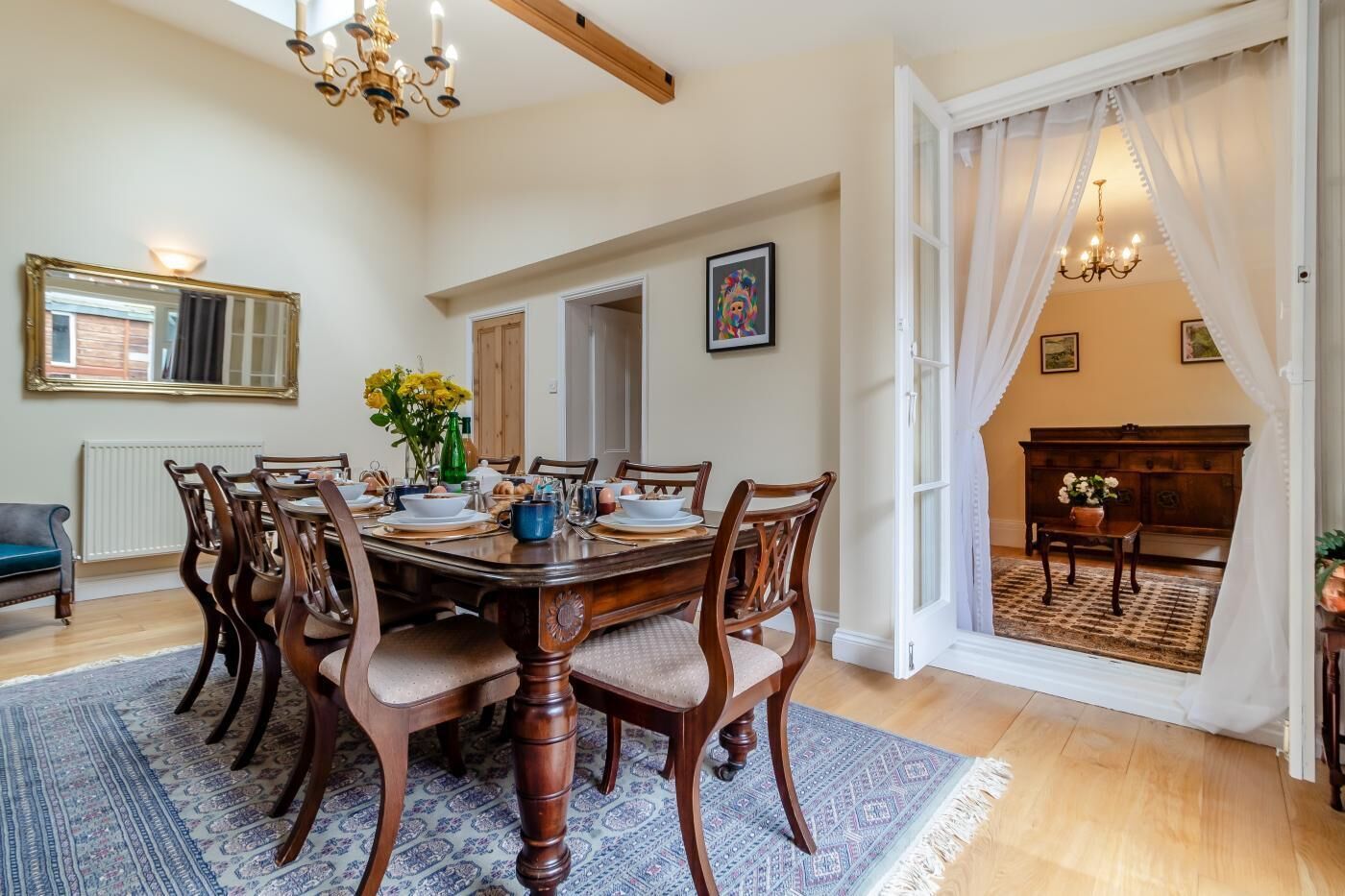 Uma sala de jantar com uma mesa de madeira posta para seis pessoas, rodeada por cadeiras. Há um espelho na parede, um candelabro e uma vista para outro espaço através de uma porta aberta.