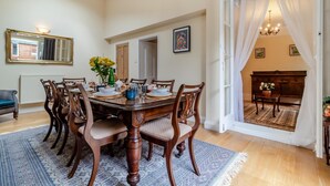 Ein Esszimmer mit einem Holztisch, gedeckt für sechs Personen, umgeben von Stühlen. An der Wand hängt ein Spiegel, und eine Kandelaber ist angebracht. Durch eine offene Tür ist ein Blick in einen anderen Raum möglich.