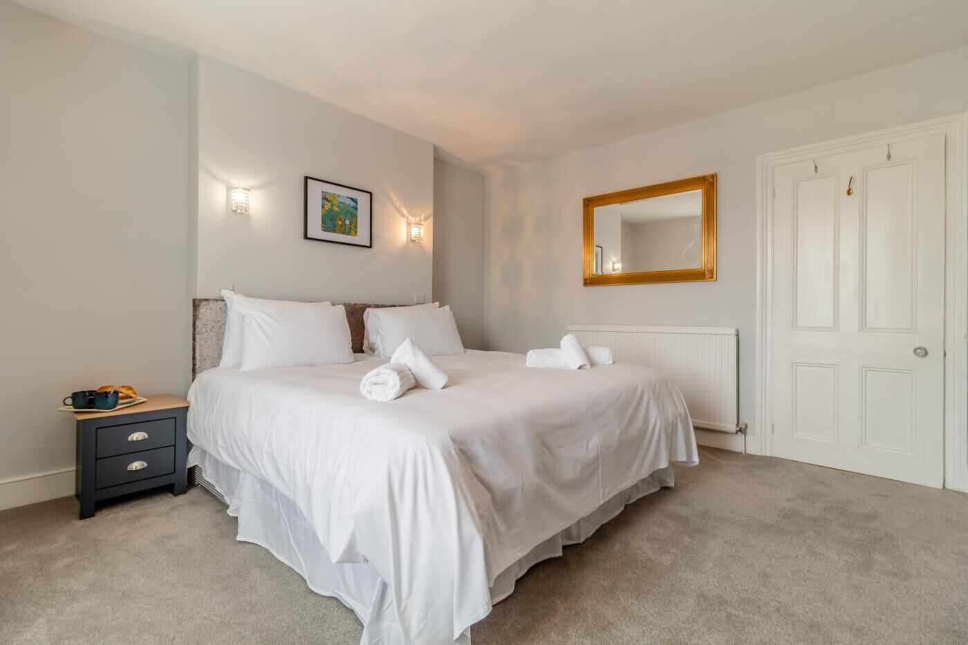 Een opgemaakt bed met witte lakens en handdoeken, een nachtkastje met een kopje en een bord, een spiegel aan de muur, en een radiator.