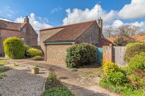 A brick house with a tiled roof, surrounded by a garden with various plants and a gravel path. - 2 Bed in Langham (oc-g25208) (Langham)