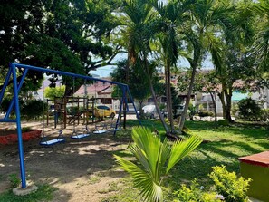 A playground with swings and a slide, surrounded by trees and a building in the background.