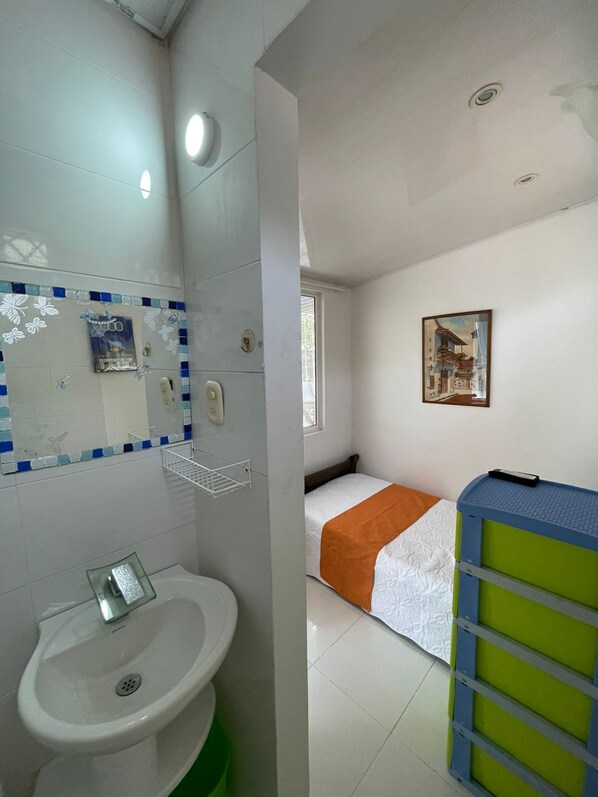 Un baño con lavamanos blanco, espejo e inodoro. Hay una cama con manta naranja y un mueble de almacenamiento verde y azul.