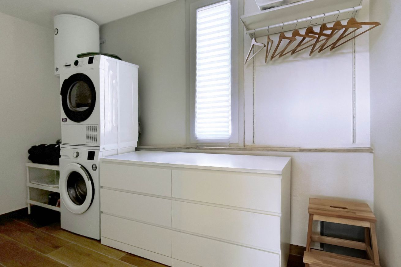 A laundry room with a stacked washer and dryer, a drying rack, and a small wooden stool.