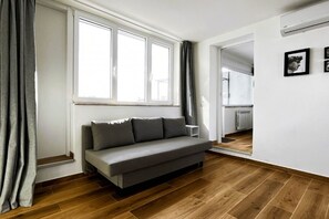 A modern living room with a grey sofa, wooden flooring, and a large window with curtains. - Alma Suites - Skyline Penthouse (Firenze)
