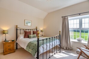 A bedroom with a four-poster bed, a wooden nightstand, a bedside table, a window with curtains, and a view of a green landscape. - 2 Bed in Pencaenewydd (oc-t29187) (Pencaenewydd)