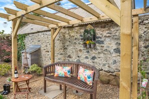 Una pérgola de madera con un banco y una mesa, un muro de piedra y una maceta colgante.