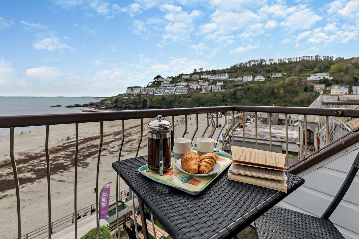 A balcony with a table set for breakfast, overlooking a beach and houses in the background.