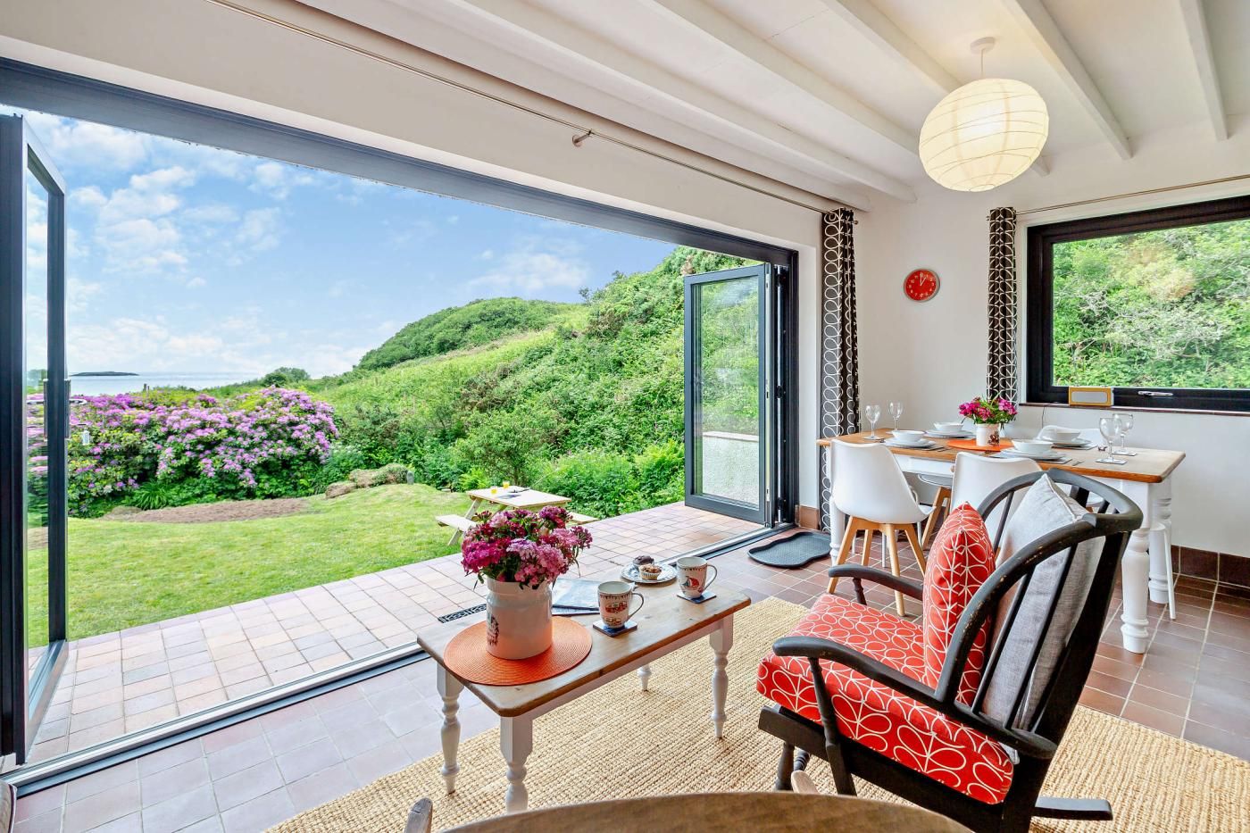 A spacious room with a large sliding glass door leading to a garden. There is a dining area with a table set for two, a sofa, and a chair.