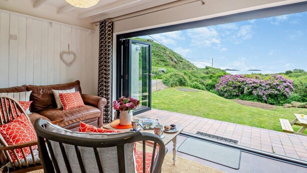 Ein modernes Wohnzimmer mit einem braunen Ledersofa, einem Holzstuhl und einem Couchtisch. Eine große Glasdoor führt in einen Garten mit Blick auf Hügel und violette Blumen.