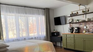 A hotel room with a bed, a dining table, a sofa, a kitchen area with a TV, and a brick wall.