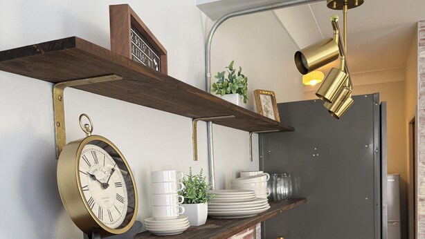 A kitchen with a brick wall, wooden shelves, a coffee maker, a sink, and a clock.