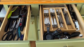 A kitchen counter with two coffee makers, a coffee pot, and a sign indicating no smoking or vaping. Below, there are two drawers with various kitchen tools and utensils.