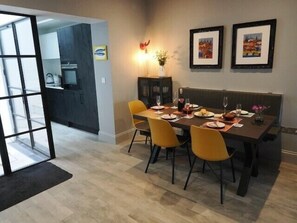 Een eetgedeelte met een donker houten tafel voor vier personen, gele stoelen en een donker houten bankje. Er hangen afbeeldingen in lijsten aan de muur, een vaas met bloemen en een klein kastje.