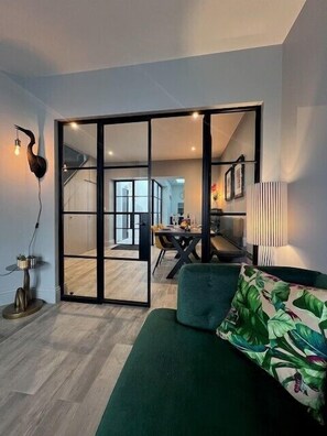 A modern living room with a green sofa, a dining area with a table and chairs, and a kitchen in the background. - 3 Bed in Fowey (oc-p29280) (Fowey)