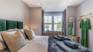 A modern hotel room with a large bed, a bedside table, a mirror, and a view of the city through large windows. - 3 Bed in Fowey (oc-p29280) (Fowey)