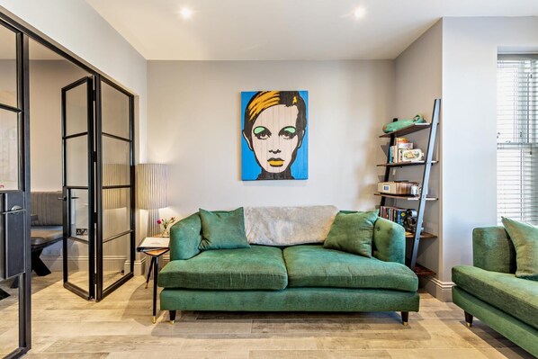 Een woonkamer met een groene bank, een schilderij aan de muur en een boekenplank.
