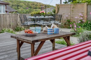 A wooden deck with a table set for two, featuring a bowl of fruit, two glasses, and a book. - 3 Bed in Golant (oc-o29307) (Golant)