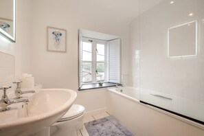 A bathroom with a white sink, toilet, and bathtub. There is a window with shutters, a framed picture on the wall, and a small window ledge with a cup. - 3 Bed in Golant (oc-o29307) (Golant)
